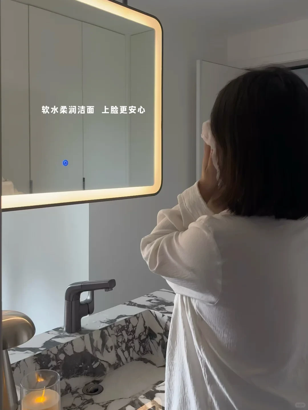 A person in a white top cleanses their face in front of an illuminated rectangular mirror above a marble bathroom vanity.
