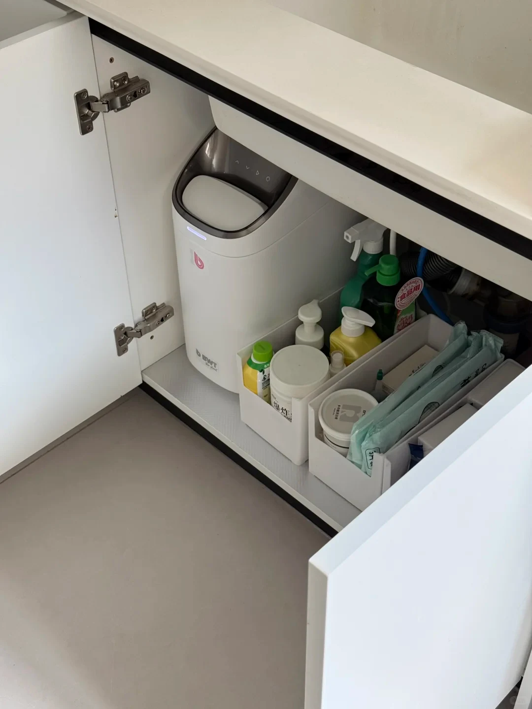 An open white under-sink cabinet containing a white BWT appliance on the left and two white organizer bins filled with various cleaning bottles and supplies on the right.