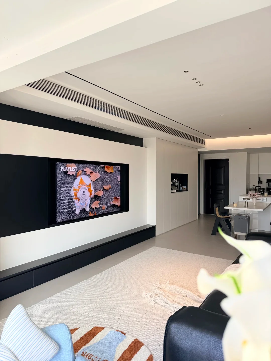 Modern minimalist living room featuring a built-in television displaying a music playlist on a black-and-white feature wall, a light-colored area rug, and an adjoining dining and kitchen area.