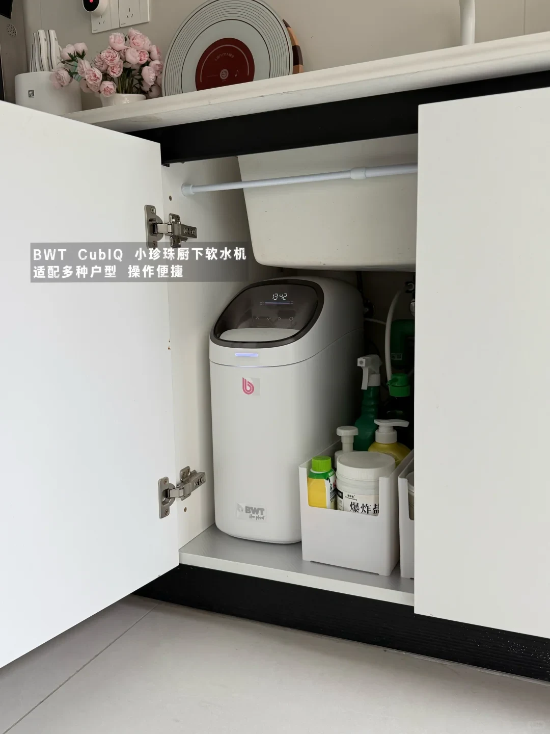 A white BWT CubIQ compact under-sink water softener installed inside a kitchen cabinet alongside household cleaning supplies.