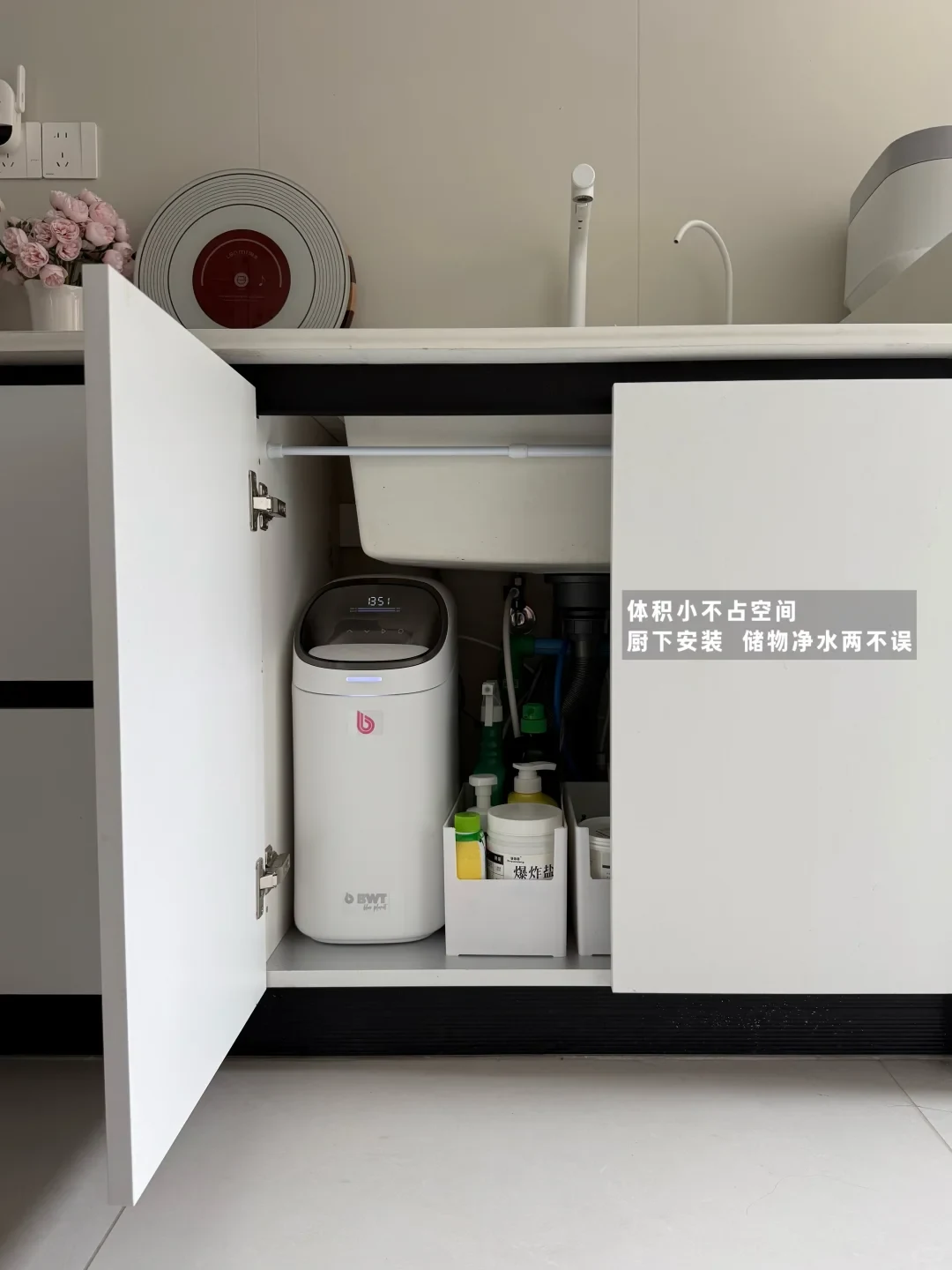 A white BWT water purifier installed inside an open under-sink kitchen cabinet beside storage bins with cleaning supplies.