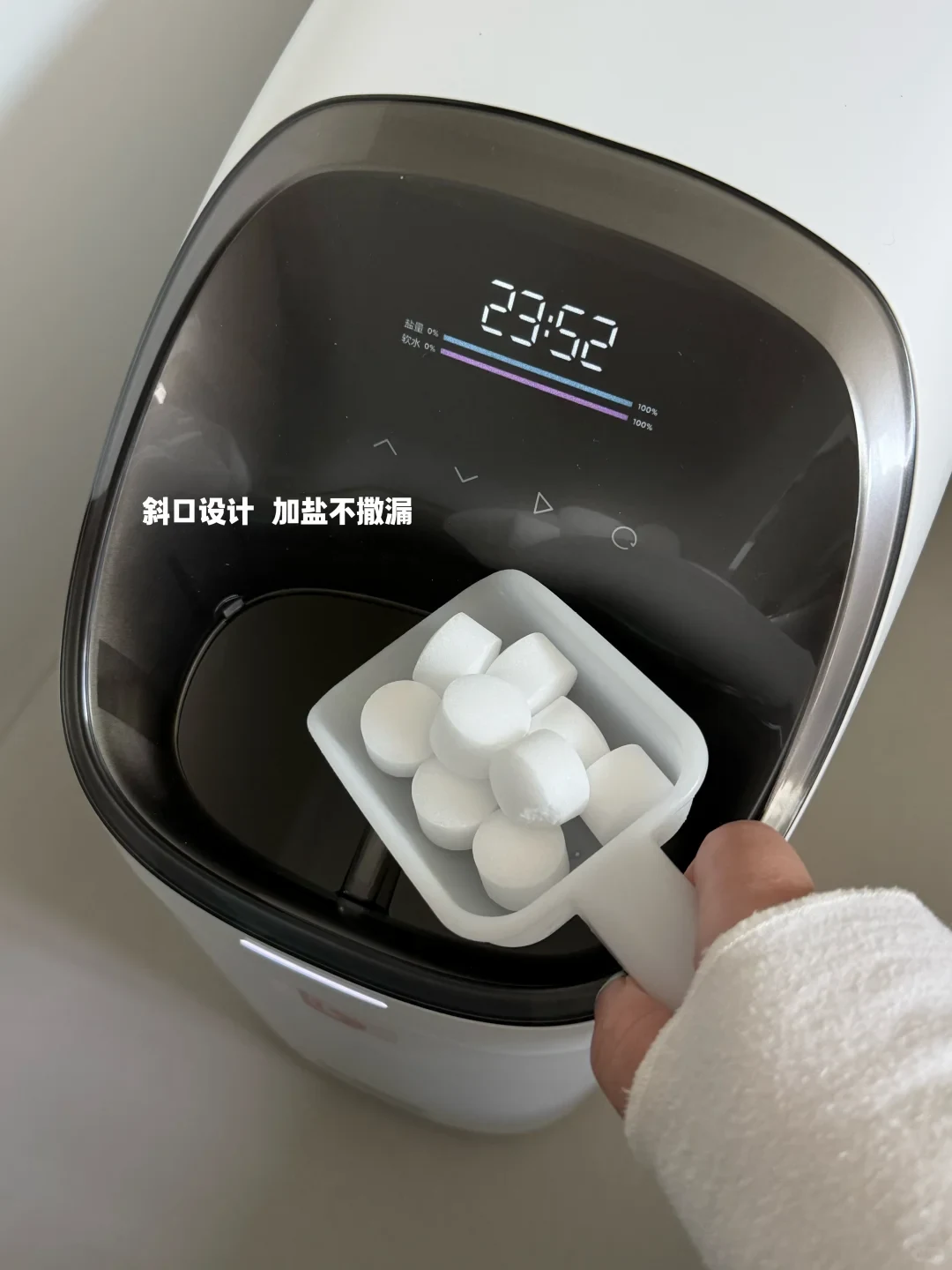 A person holding a scoop of white salt tablets over the open salt tank of a compact white water softener with a digital display.
