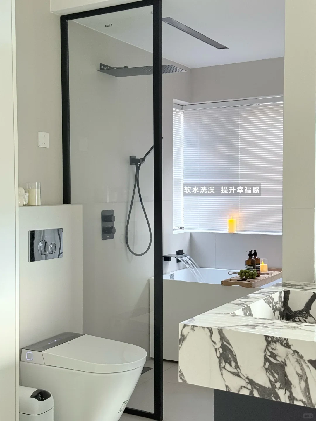 Modern bright bathroom featuring a white smart toilet, a glass-enclosed shower area with black hardware, and a bathtub with water pouring from a waterfall spout beneath a window with blinds.