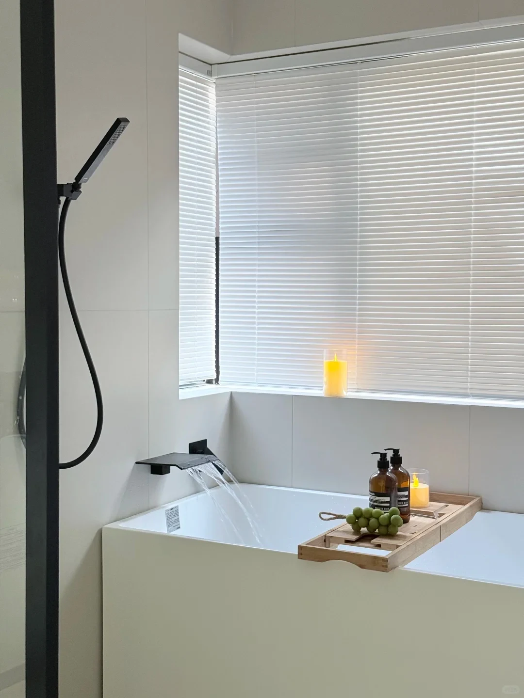 A minimalist modern white bathtub filling with water from a matte black waterfall faucet, with a wooden bath caddy holding bottles, a candle, and green grapes beneath a window with white blinds.