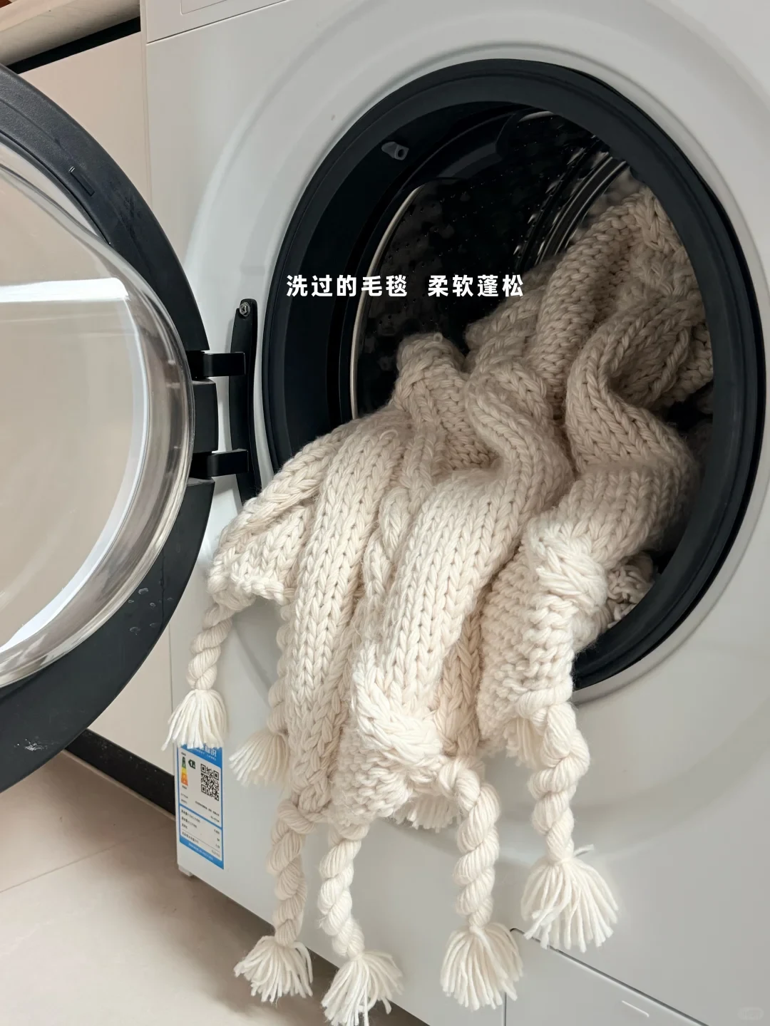 A chunky cream-colored knitted blanket with braided tassel fringe draped out of an open front-loading washing machine drum.