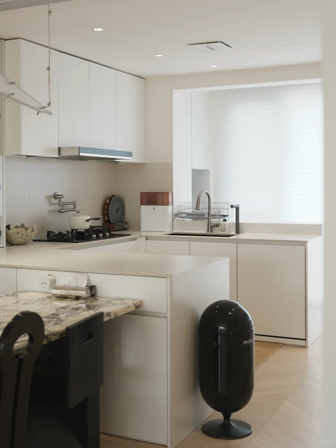 A modern minimalist kitchen interior featuring white cabinetry, a kitchen island counter attached to a marble dining table, a sink by a large window, a gas stovetop with wall-mounted pot filler, and a black pedestal capsule trash can on the wooden floor.
