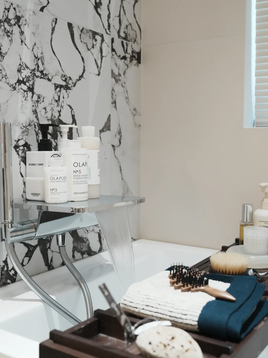 A wide waterfall bathtub faucet filling a white tub, with toiletries including Olaplex bottles arranged on its chrome shelf surface, and a wooden bath caddy holding brushes and folded towels in the foreground against a marble wall.