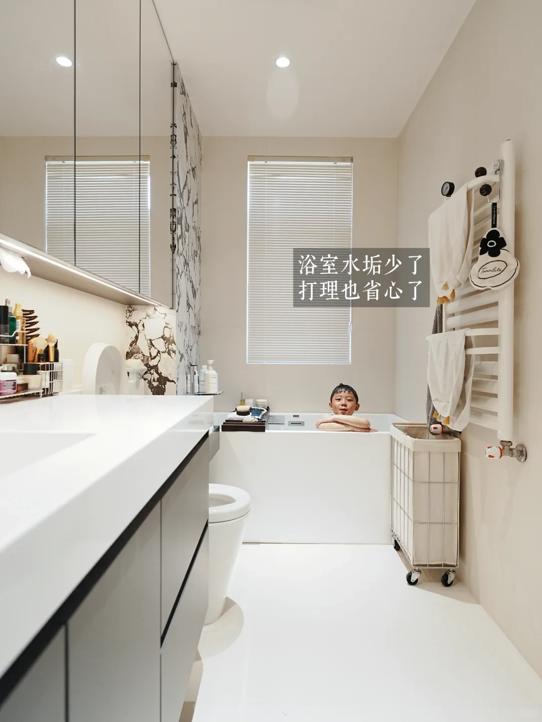 A child relaxes in a bathtub at the far end of a sleek, modern white bathroom featuring a vanity counter, mirrored cabinets, and a heated towel rack.