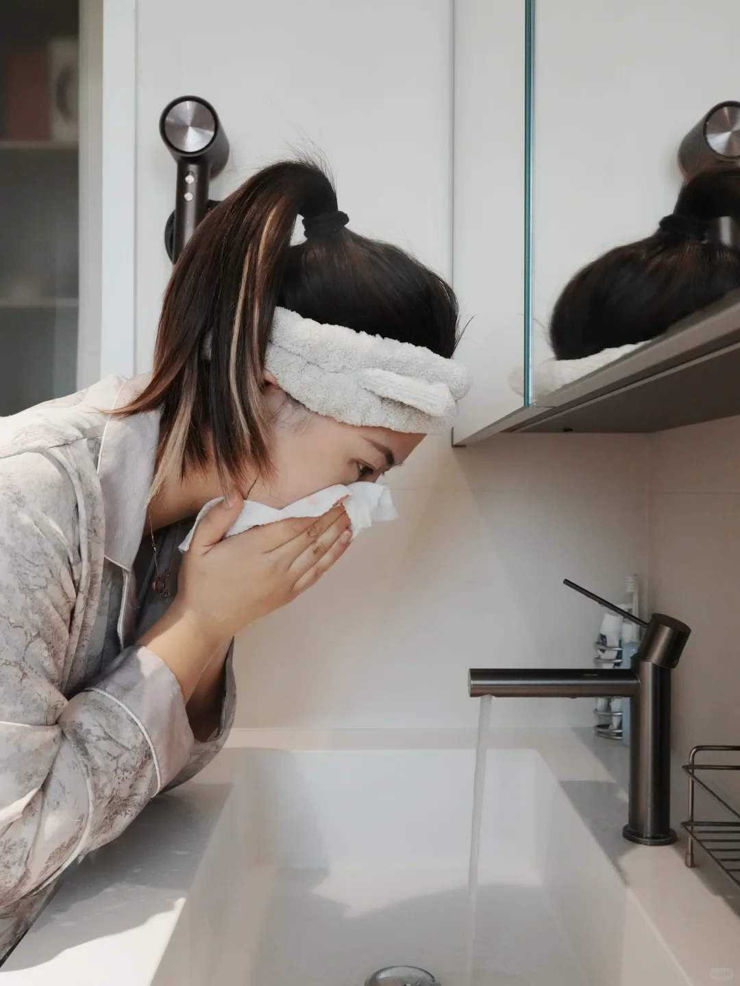 A woman wearing a headband bends over a bathroom sink, using a disposable face towel to wipe her face as water flows from the modern faucet.