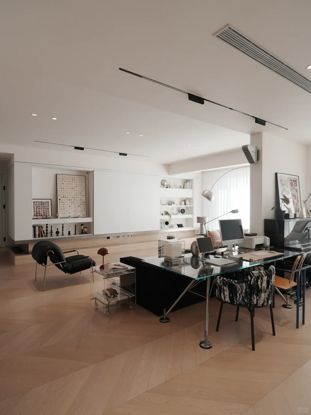 Spacious modern living room and home office featuring light wood chevron flooring, a glass-top desk with a desktop computer and chairs in the foreground, a black leather lounge chair, and a minimalist white built-in wall unit with display shelves and art prints in the background.