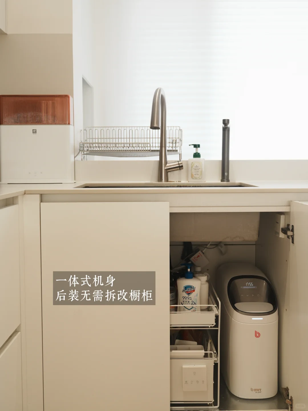 A kitchen sink setup showing the open under-sink cabinet containing a compact BWT water softener and organized pull-out storage shelves.