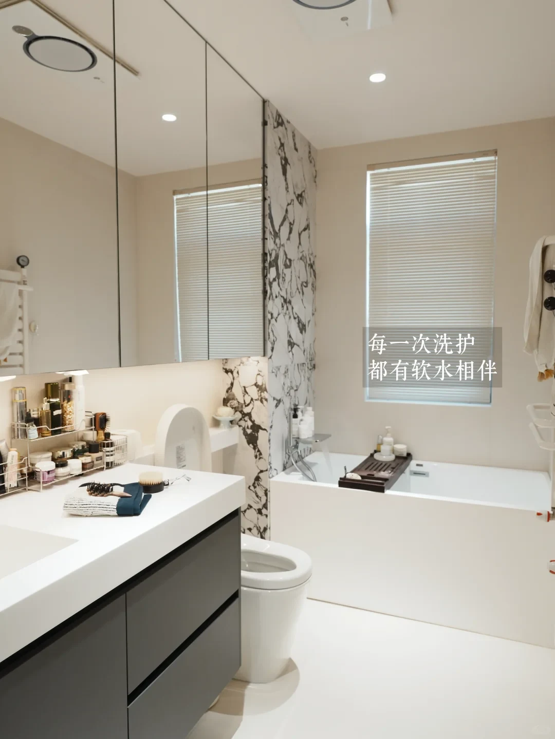 Bright modern bathroom with a mirror cabinet above a vanity with skincare products, a toilet, and a bathtub with a bath tray under a window with blinds.