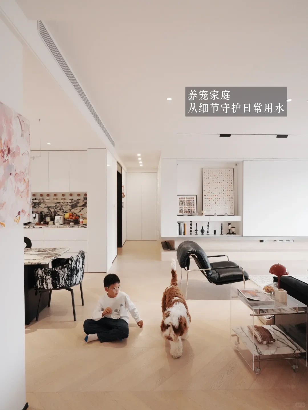 A modern bright living room where a young boy sits on the wooden floor holding a cup of water beside a walking dog.