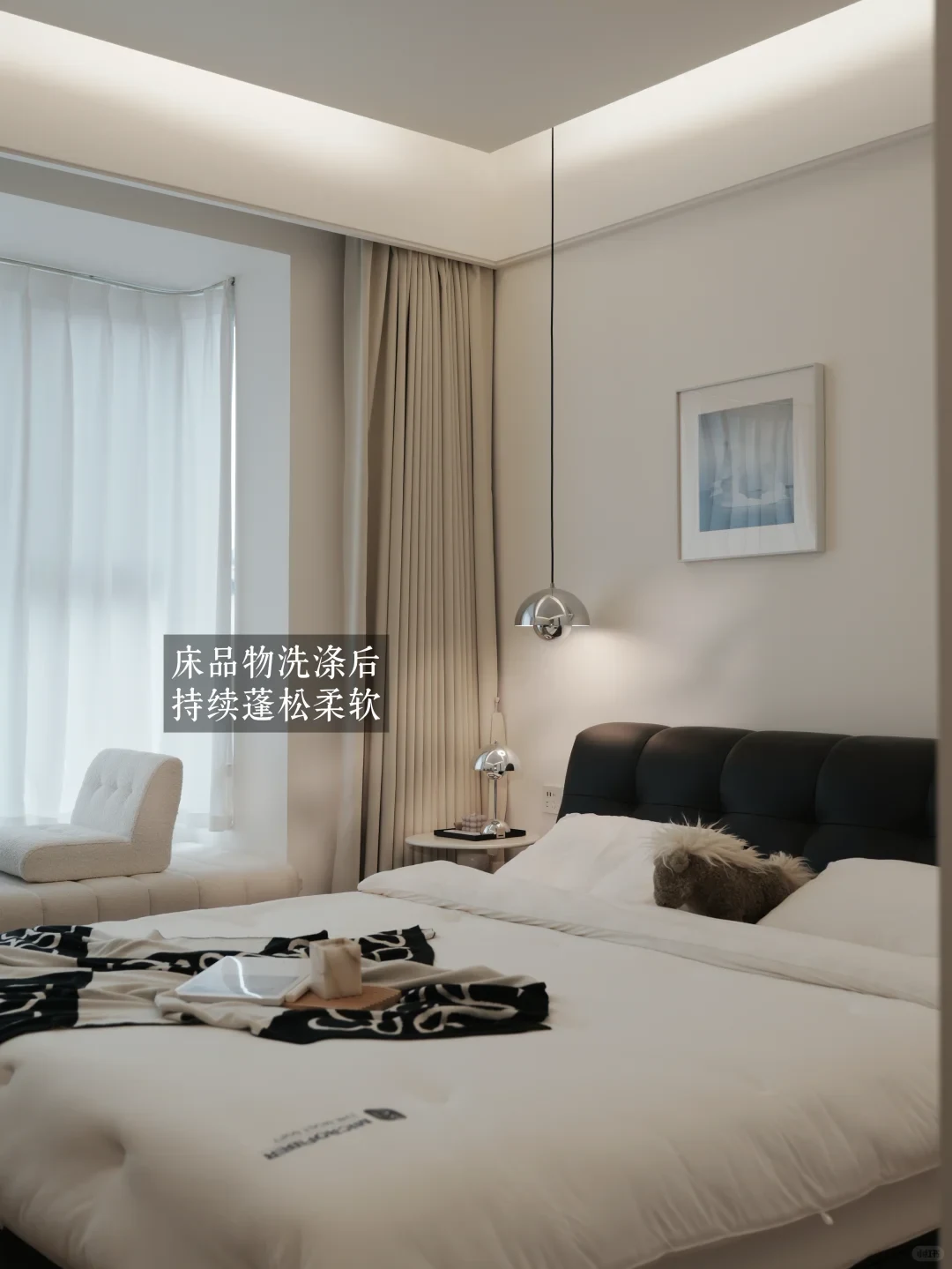 A modern minimalist bedroom interior with a neatly made bed dressed in fluffy white bedding, a black tufted headboard, and a chrome pendant light hanging near the curtains.