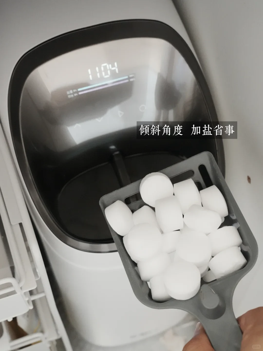 A hand holds a gray scoop filled with round white salt pellets over the open salt tank of a modern white water softener machine featuring a digital display.
