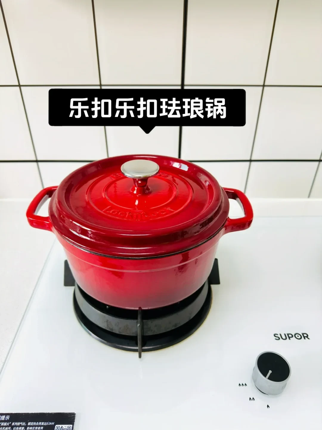 A red LocknLock enameled cast-iron pot with lid set on a white gas stovetop against a tiled wall.
