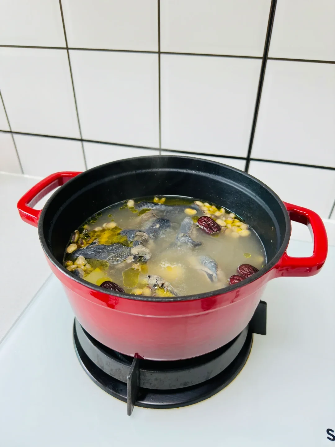 A red cooking pot on a stove burner filled with simmering black chicken soup containing jujubes and beans.