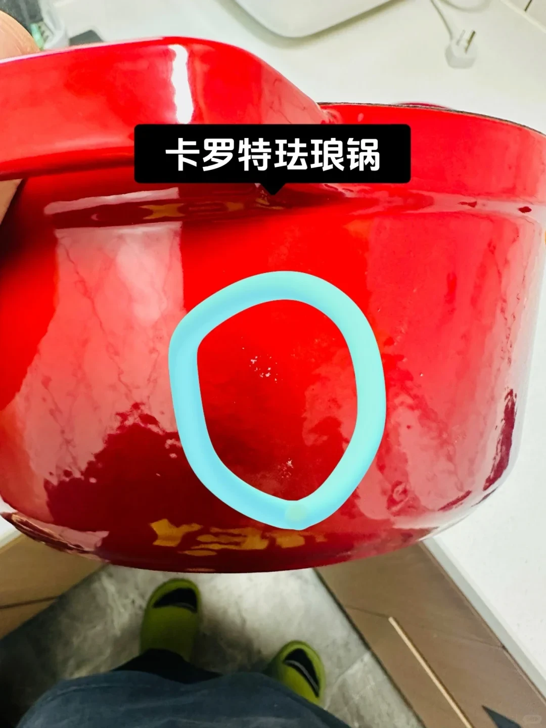 A glossy red enamel Dutch oven held in hand, with a blue ring drawn around small specks on its side and a black label tag above.