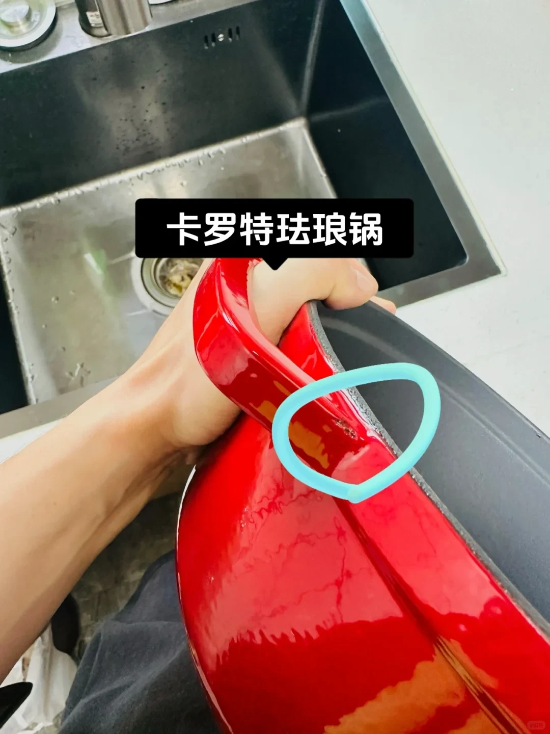 A person holds the handle of a red Carote enameled cast iron pot over a sink, showing a chipped enamel defect on the rim circled in light blue.