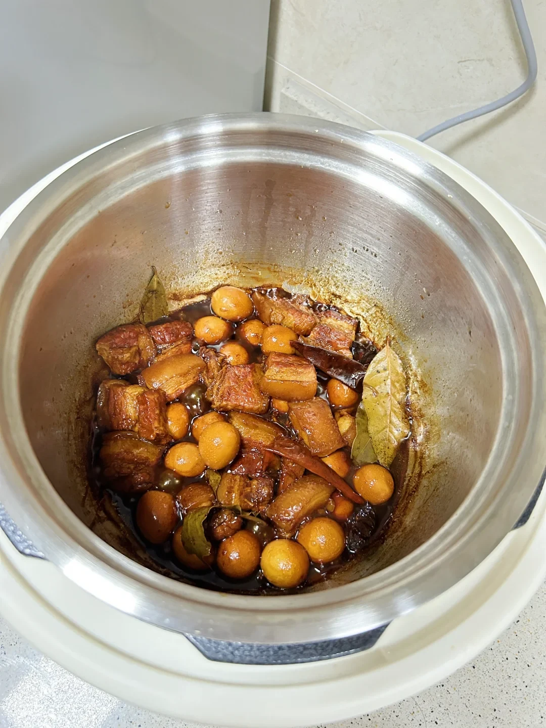 Top-down view of braised pork belly and quail eggs cooked in a glossy dark sauce with bay leaves inside the stainless steel inner pot of an electric pressure cooker.