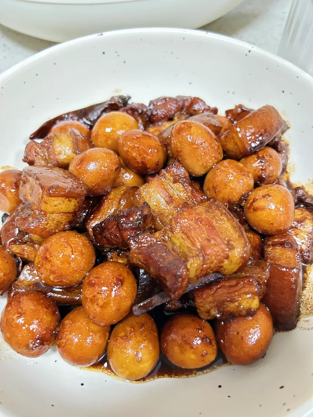 A white ceramic bowl filled with glossy braised pork belly pieces and whole braised quail eggs coated in a rich, dark brown glaze.
