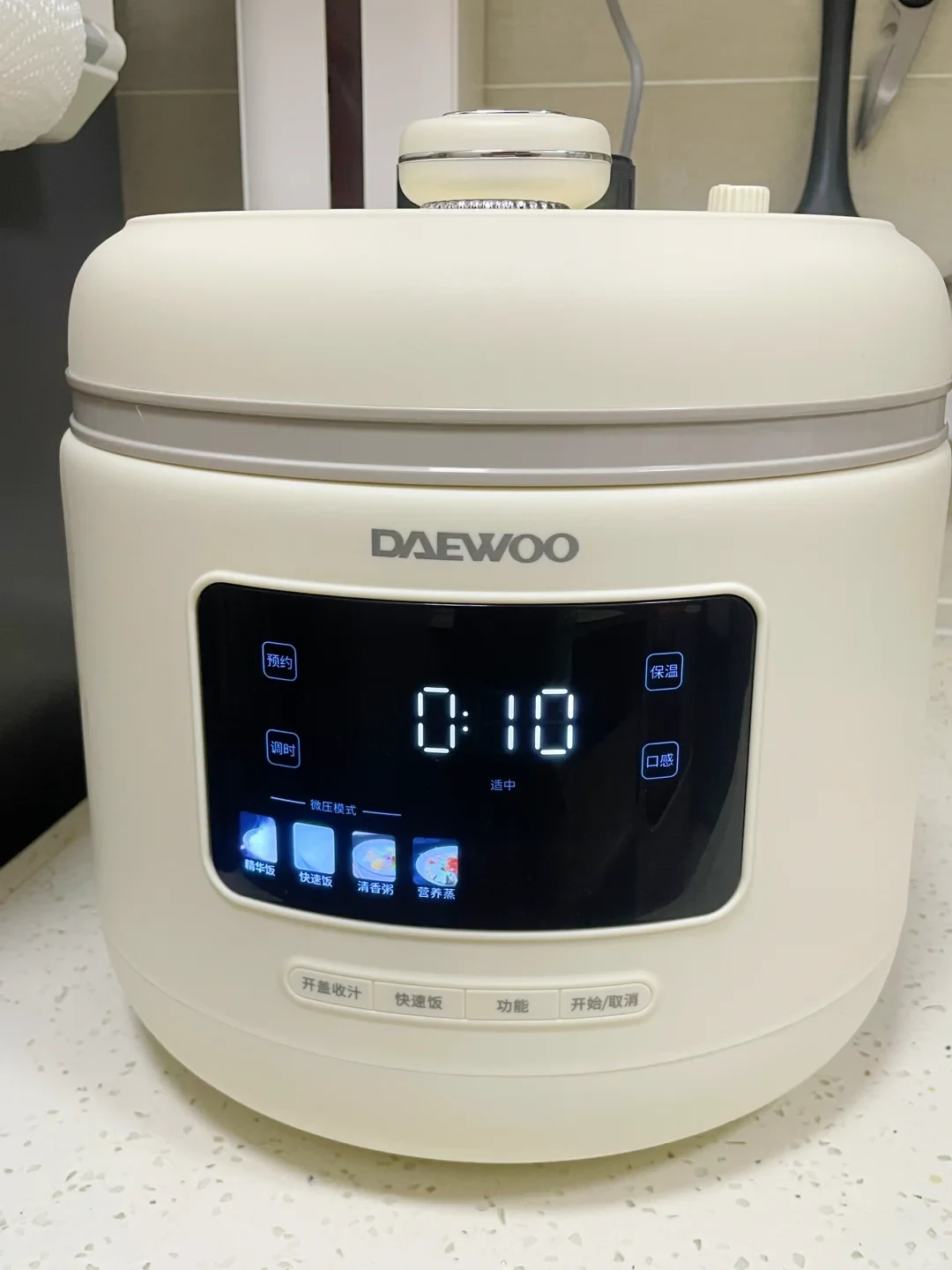 A white Daewoo electric multi-cooker sitting on a kitchen counter with its digital display lit up with a timer reading 0:10.