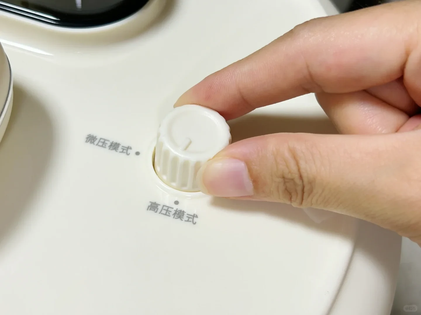 A person's hand turning a white knob on a white pressure cooker, with mode markings printed next to it.
