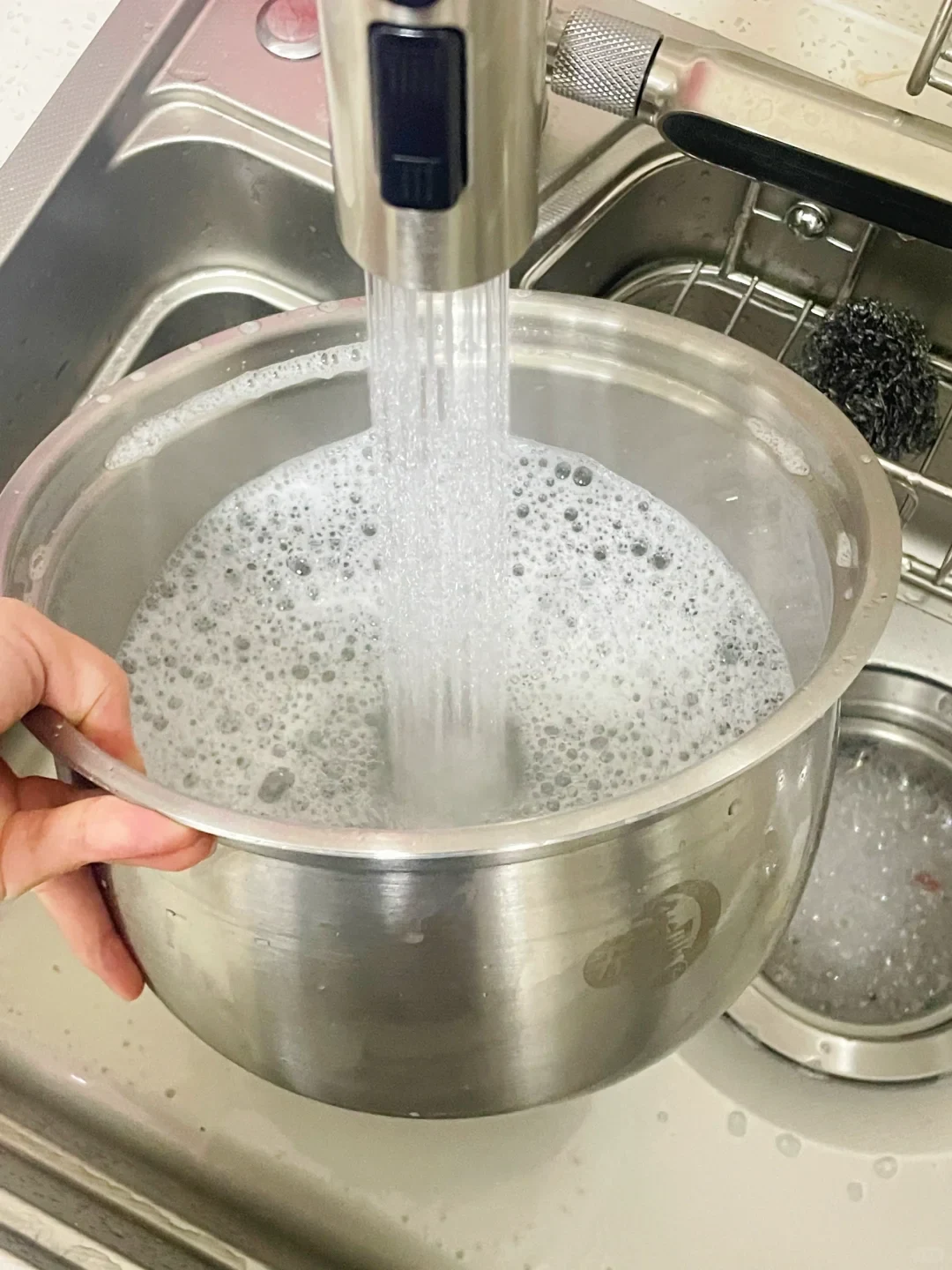 A hand holds a stainless steel mixing pot under a kitchen faucet sprayer, filling it with foamy, soapy water inside a stainless steel sink.