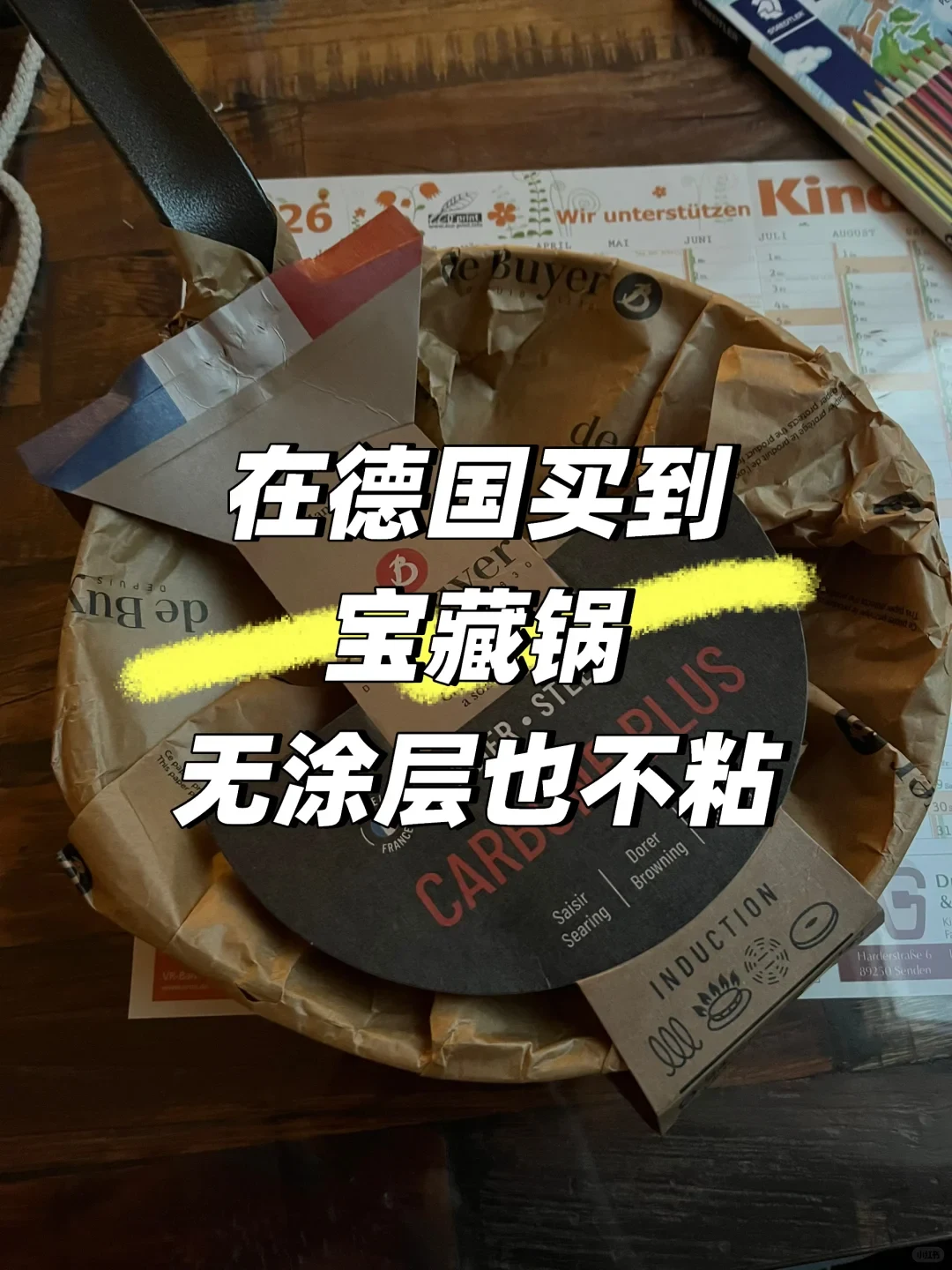 A de Buyer carbon steel frying pan sitting in brown paper packaging on a table next to a German calendar, with large Chinese text overlaid in the center.