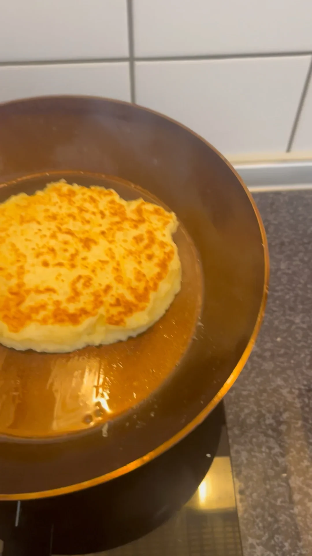 A thick, golden-brown pancake or potato cake frying in oil inside a round skillet on a stovetop.