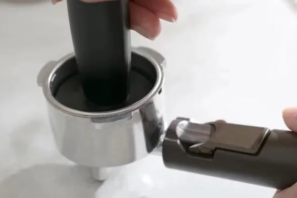 A hand presses coffee grounds down into a stainless steel portafilter basket using a black tamper.