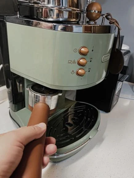 A hand holding the portafilter handle into the group head of an olive green De'Longhi ECO310 espresso machine.