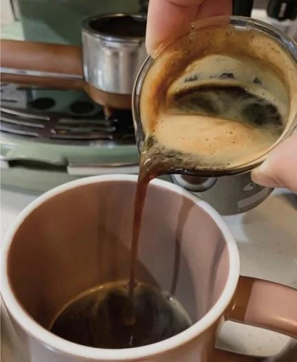 Freshly extracted espresso with a thick layer of crema being poured from a glass measuring pitcher into a ceramic mug, with the portafilter and De'Longhi espresso machine visible in the background.