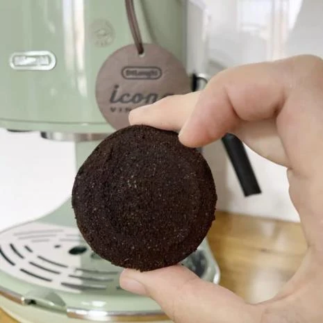 A hand holding up a round compacted espresso puck in front of a pale green De'Longhi Icona coffee machine.