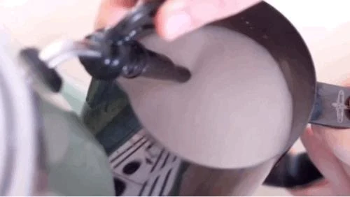 Top-down view of a steam wand immersed in milk inside a stainless steel frothing pitcher.