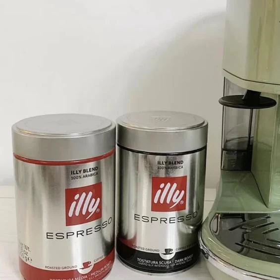 Two tin cans of illy espresso ground coffee standing next to a vintage green De'Longhi espresso machine on a counter.