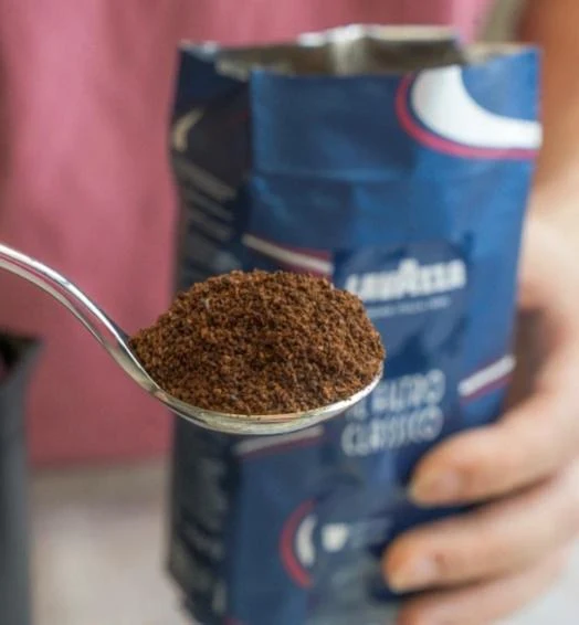 A metal spoon heaped with finely ground coffee held in front of an open blue bag of Lavazza coffee.