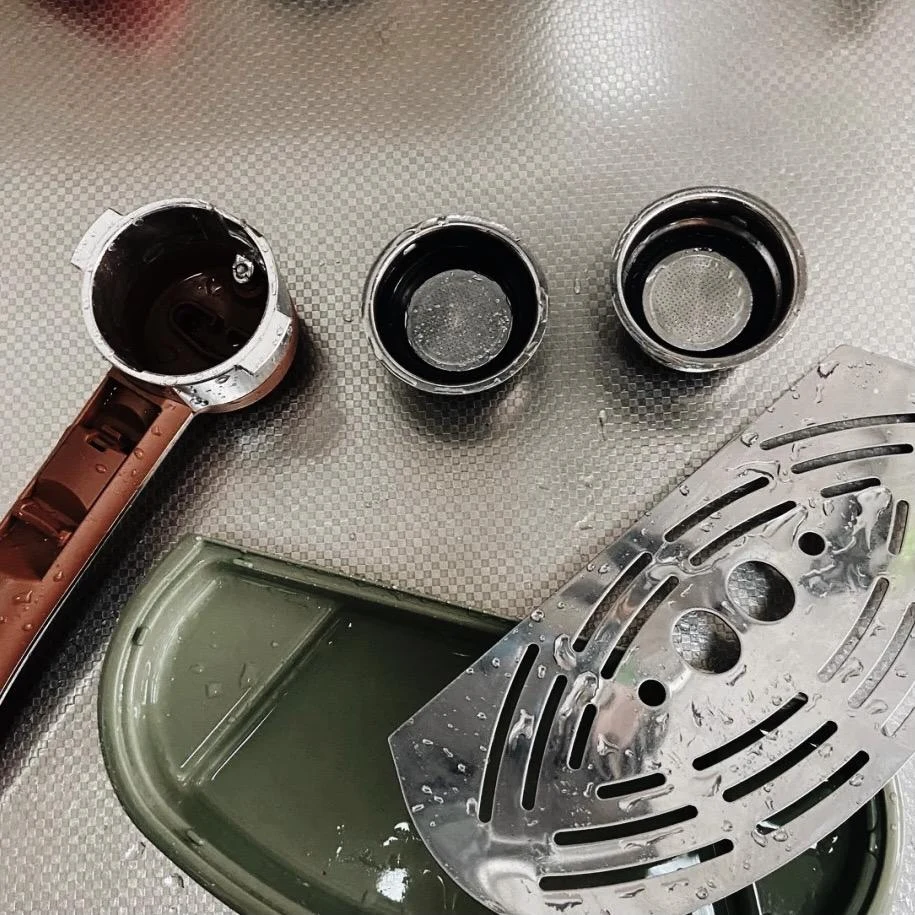 Top-down view of disassembled and freshly washed coffee machine parts, including a portafilter handle, two round filter baskets, an olive green drip tray, and a stainless steel drip grid on a textured countertop.