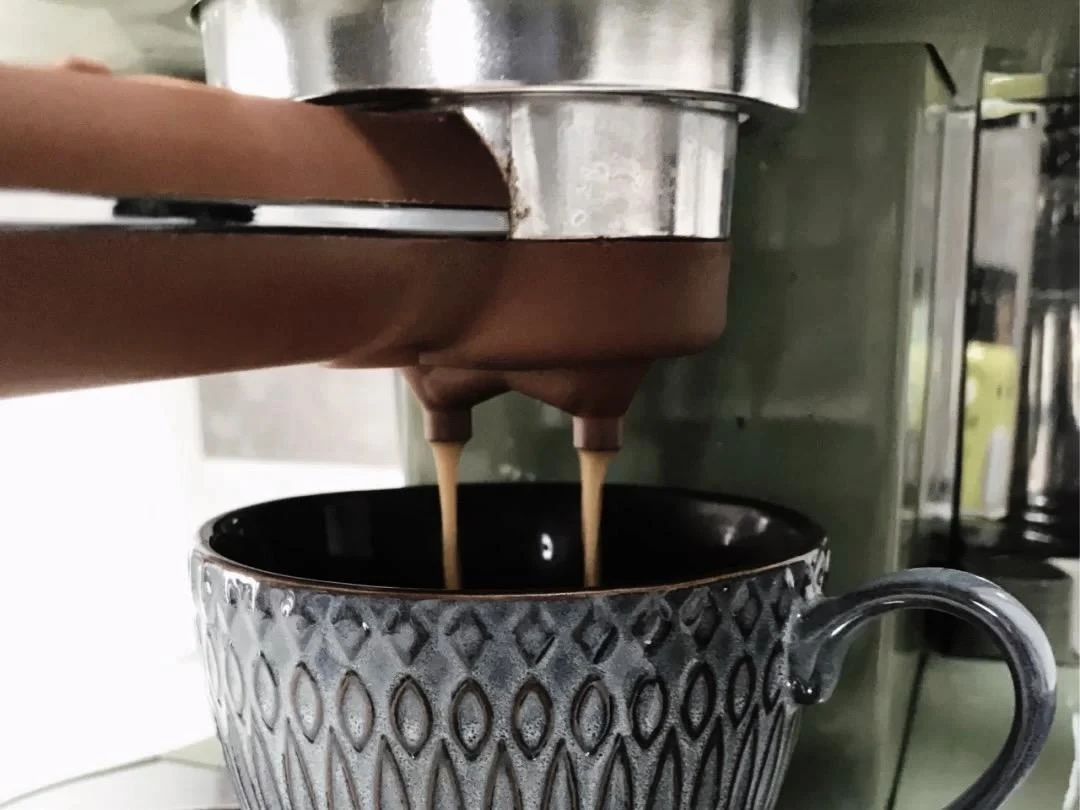 Close-up of espresso flowing from the dual spouts of a portafilter on an olive green coffee machine into a patterned ceramic cup.