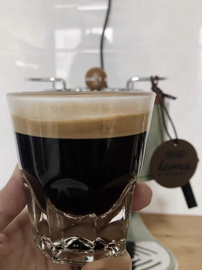 A hand holding a clear faceted glass tumbler filled with freshly brewed dark espresso topped with thick golden crema, with an olive green De'Longhi Icona espresso machine in the background.