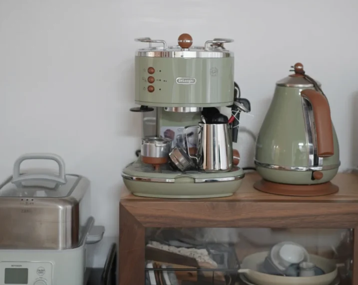 A retro pale-green De'Longhi espresso machine sits on a wooden cabinet beside a matching electric kettle, with a toaster partly visible at left.