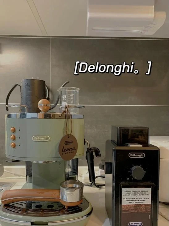 A pale green De’Longhi retro-style espresso machine sits beside a black De’Longhi coffee grinder on a kitchen counter, with a metal milk pitcher and other accessories on top.