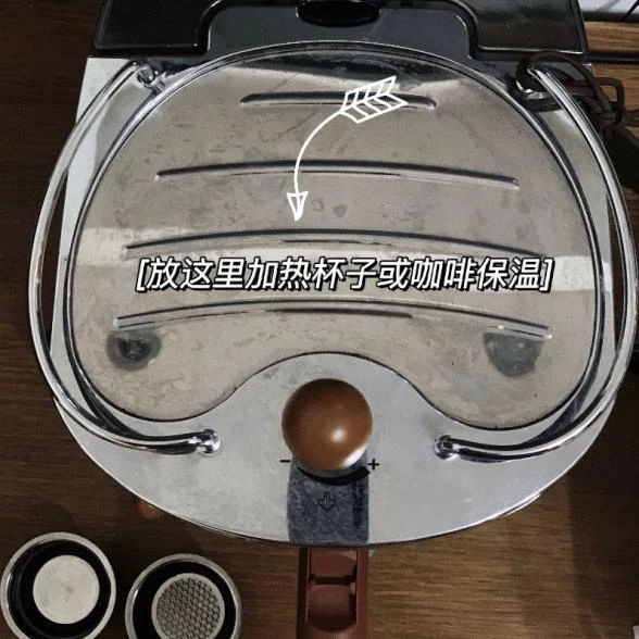 Top-down view of a compact espresso machine with a metal warming plate, a central steam-control knob marked with minus and plus symbols, and two filter baskets visible at the bottom.