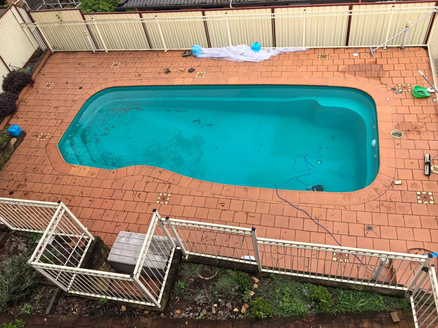 High-angle view of an outdoor backyard swimming pool with light blue water and terracotta-paved surround, showing a pool cleaner robot submerged at the bottom with a blue cable.