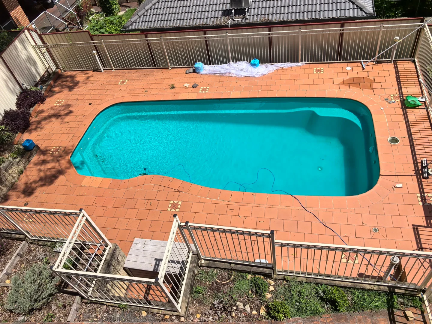 High-angle view of a fenced backyard swimming pool surrounded by terracotta tiles, showing clear turquoise water with a pool cleaner's blue power cable running into it.