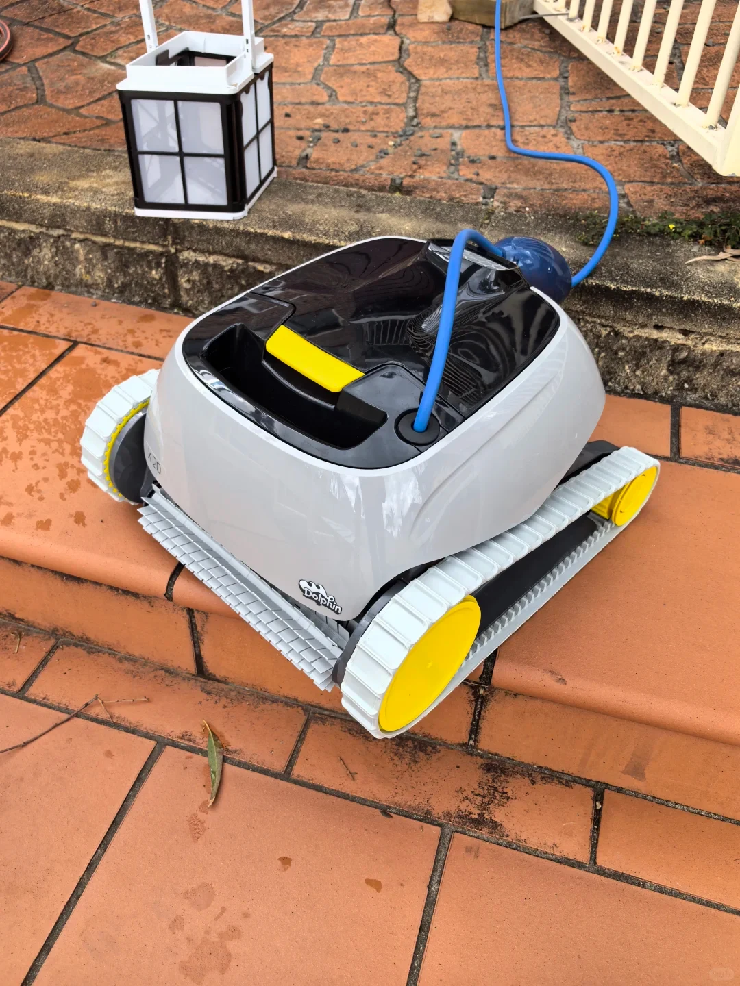 A Dolphin X20 robotic pool cleaner resting on tiled ground with its blue power cord attached and a filter basket set behind it.