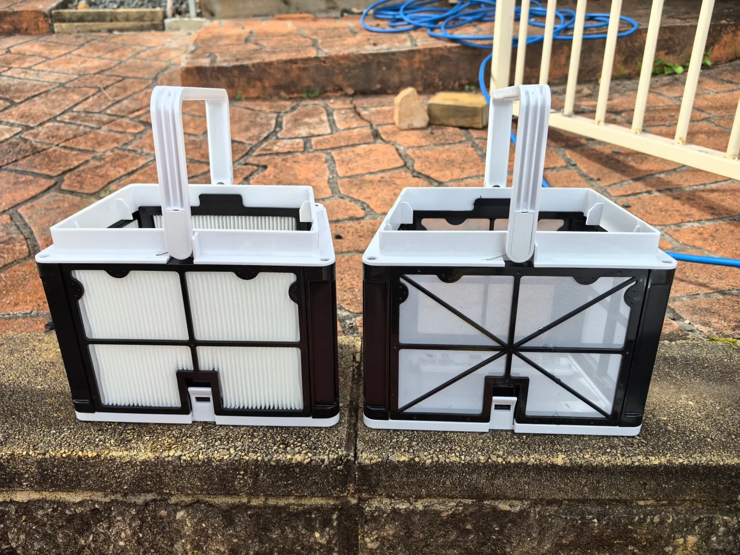 Two white and black pool cleaner filter baskets with carry handles placed side-by-side on an outdoor stone ledge, comparing a fine pleated filter paper basket on the left with a mesh filter basket on the right.
