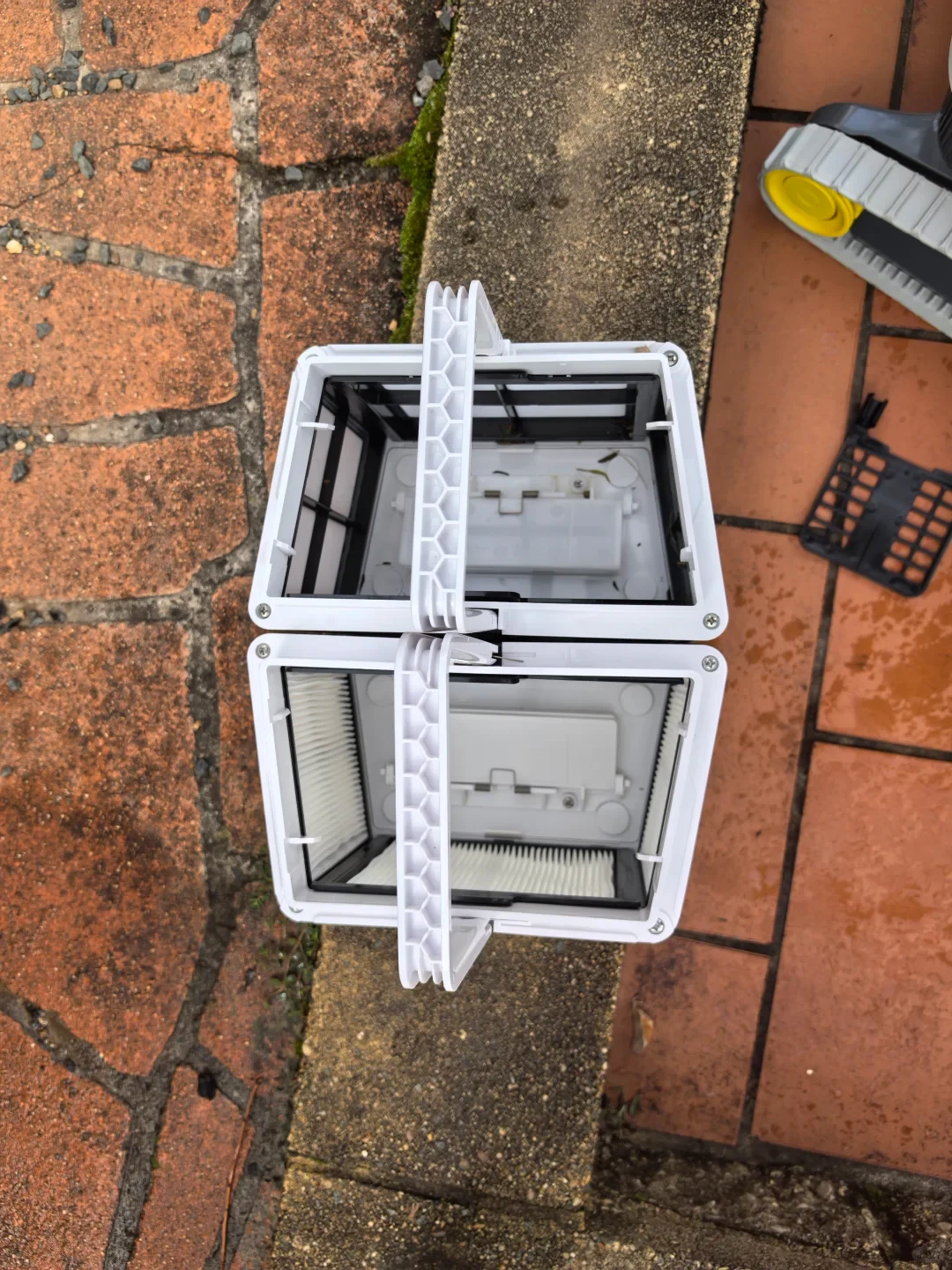 Two white pool-cleaner filter baskets are open on outdoor paving, with a standard dark mesh filter basket above and a pleated filter basket below; part of the cleaner is visible at upper right.
