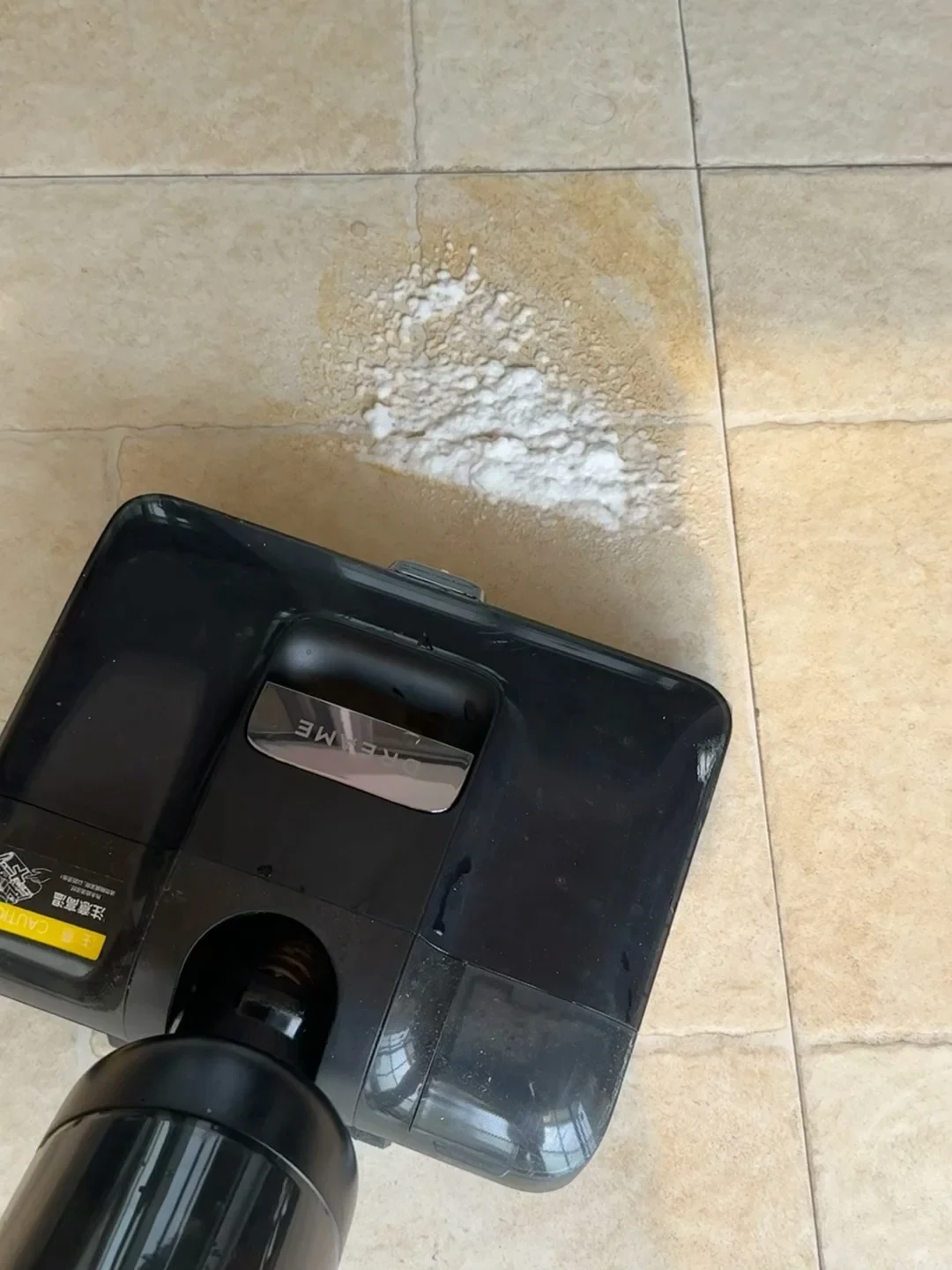 A black wet-and-dry floor cleaner approaching a spilled brown puddle topped with thick white foam on a beige tiled floor.