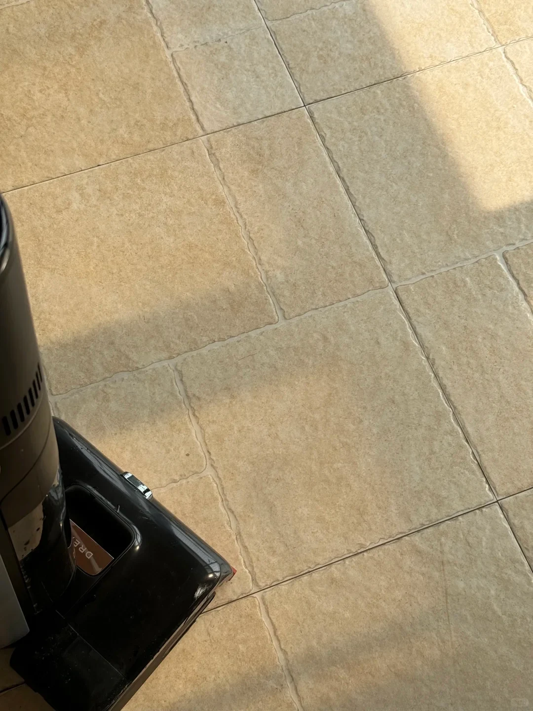A high-angle view of beige stone-textured floor tiles in sunlight, with the black base and cylinder of a vacuum cleaner or floor scrubber visible in the bottom-left corner.